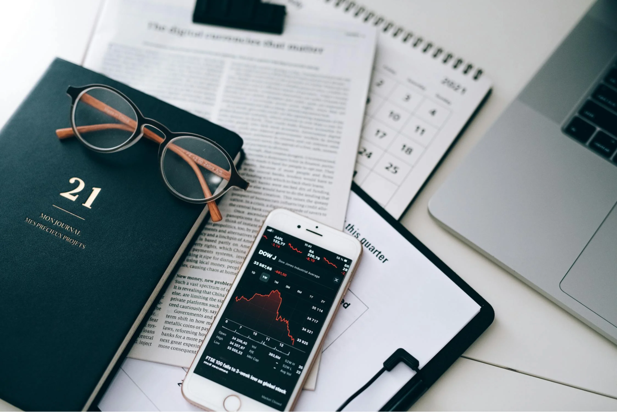 smartphone displaying a stock market chart lying on documents next to a laptop on the desk